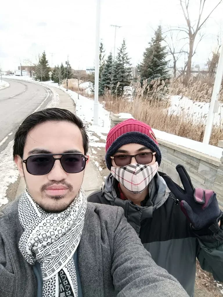 Couple wearing winter hats and face masks outdoors near road with snow and trees, casual street photo