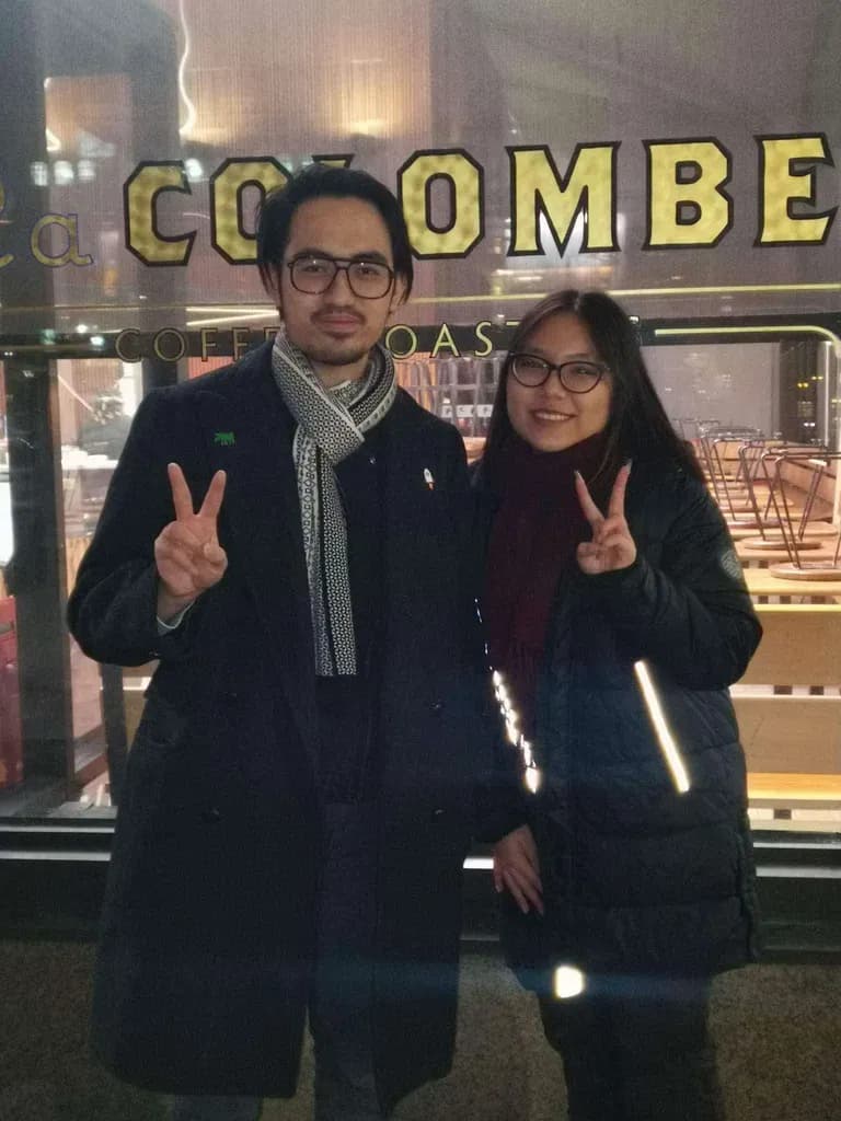 Couple posing in front of “Welcome to Combe” storefront at night, smiling and making peace signs