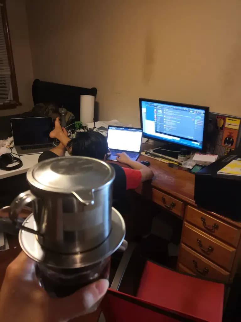 Hand holding stainless steel kettle in a home office with multiple computer monitors in background