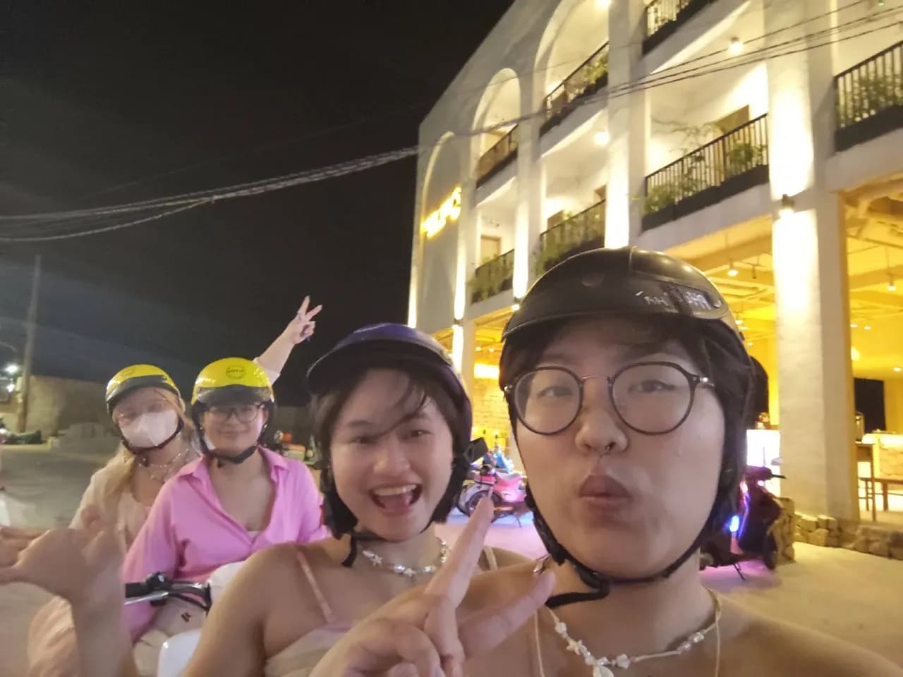 Friends wearing face masks and party costumes at night outside a lit hotel building, smiling and posing