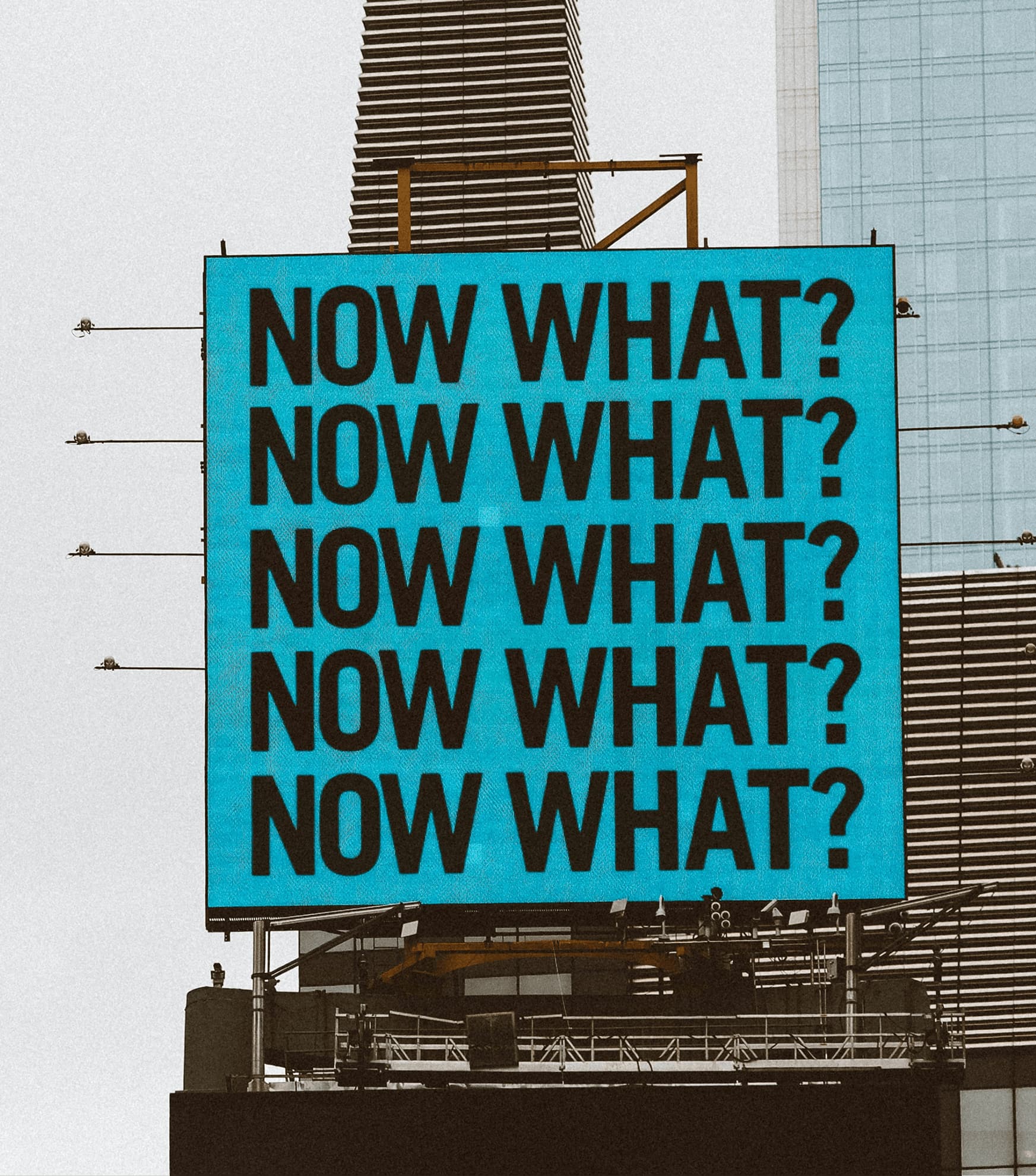 “NOW WHAT? “ repeating text on teal billboard sign in city skyline, motivational street message
